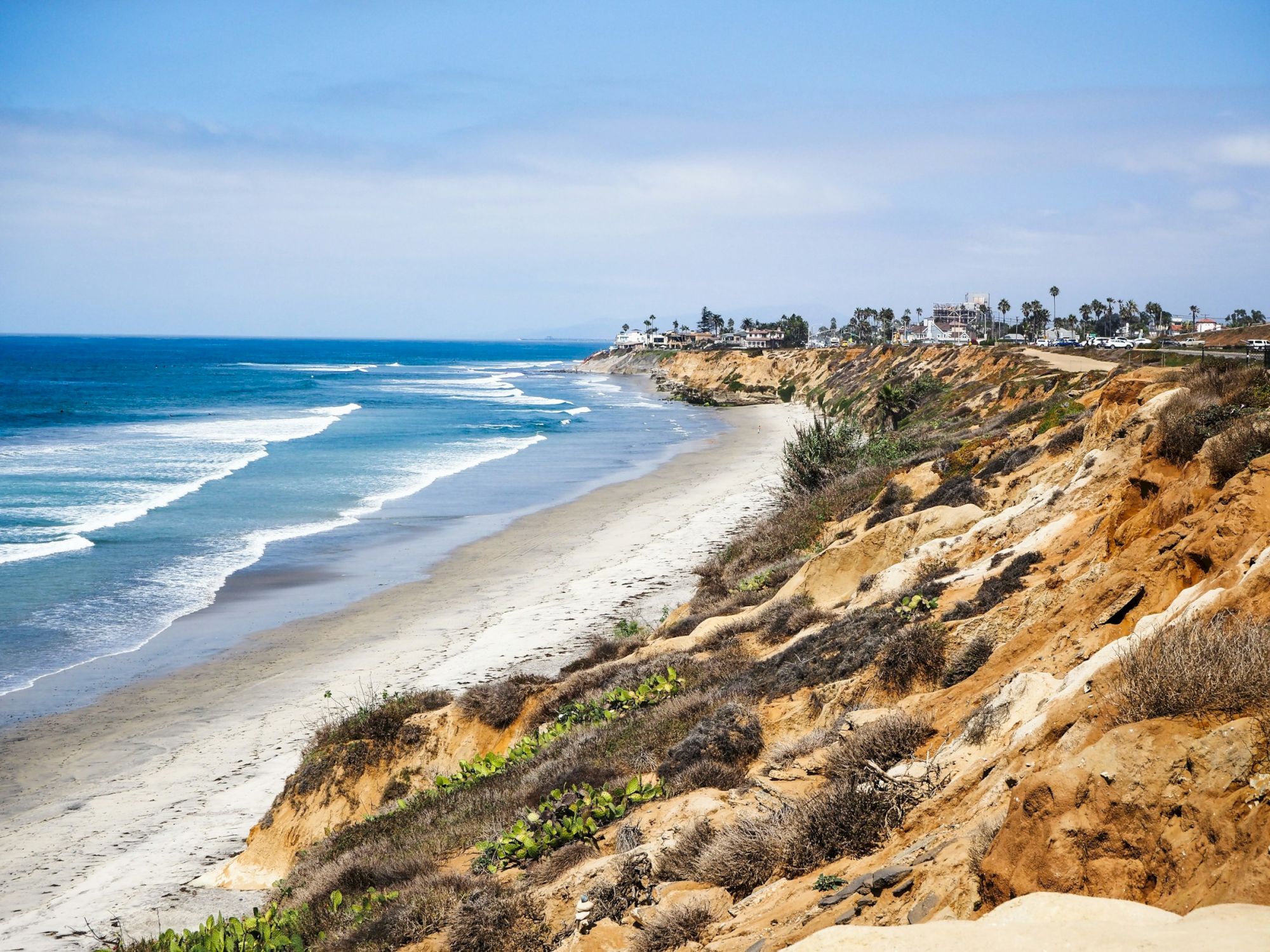 A sunny coastal scene with a sandy beach, blue ocean waves, and rocky cliffs along the shoreline, palm trees dotting the distant horizon.