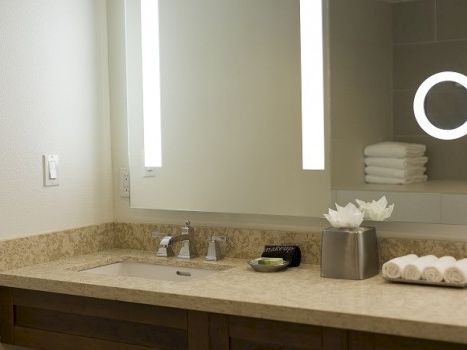 A modern bathroom vanity with beige granite countertop, a sink, stacked towels, a mirror with built-in lights, and a circular makeup light.