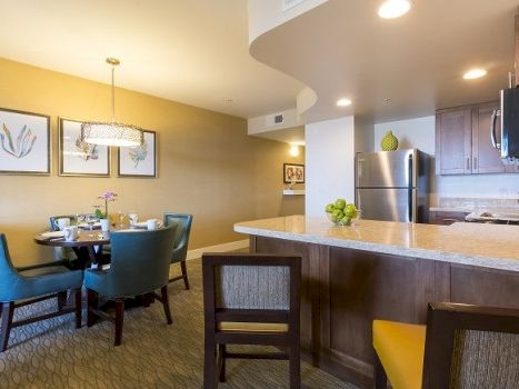 A cozy dining area and kitchen with a round table, teal chairs, warm yellow walls, framed art, and a breakfast bar with stools.