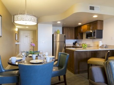 A modern open-plan kitchen and dining area with a round table, blue chairs, stainless steel appliances, and a cozy seating nook in warm lighting.