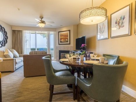 A cozy hotel suite living and dining area with a round table, eight chairs, a sofa, coffee table, wall art, and a balcony view.