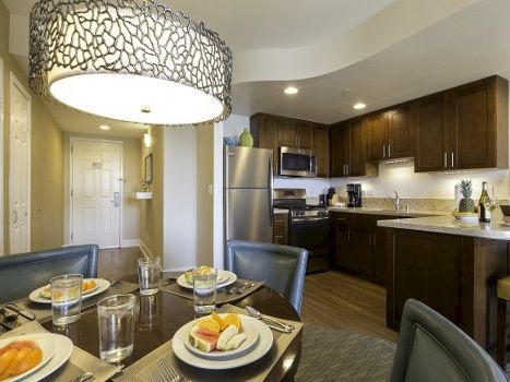 A modern kitchen-dining area with dark wood cabinets, stainless steel appliances, a circular pendant light, and a small table set for four.