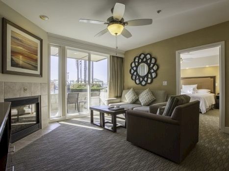 A modern hotel living room with a fireplace, large glass door to a balcony, sofa set, coffee table, decorative mirror, and a doorway to a bedroom.