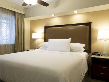 A neatly made hotel bed with white linens, two bedside tables, lamps, a window with curtains, and a ceiling fan above.