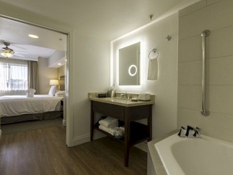 A modern hotel bathroom with a tub, vanity with sink, backlit mirror, and an open doorway to a bedroom in the background.