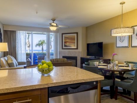 A modern living space with a kitchen island, cozy seating, and a dining area; sliding doors open to a balcony with palm trees outside.