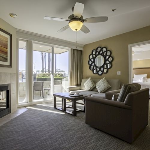A hotel suite living area with a fireplace, sofa set, coffee table, wall mirror, balcony doors, ceiling fan, and a doorway to a bedroom.