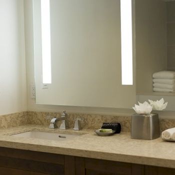 A modern bathroom sink area with a beige granite counter, mirror with vertical lights, towels, and a circular illuminated mirror in the corner.