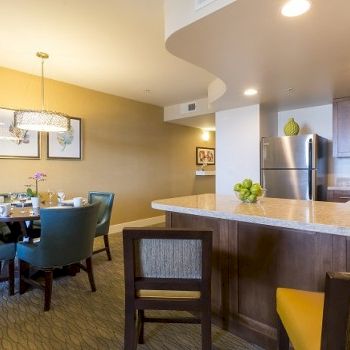 A modern kitchen and dining area with a beige wall, round dining table, teal chairs, a breakfast bar, and stainless steel appliances under warm lighting.