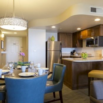 A modern apartment kitchen and dining area with a round table, blue chairs, pendant light, stainless fridge, and wooden cabinets; cozy and stylish.