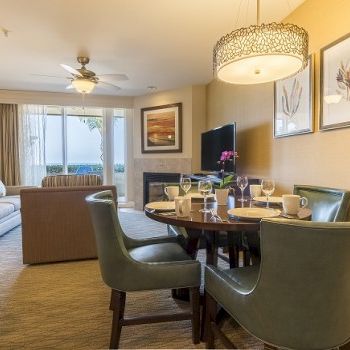 Cozy hotel suite with a dining area, a round table and four green chairs, living room in background, warm lighting, retreat vibe.