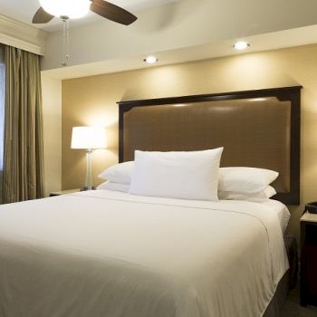 A hotel bedroom with a large bed, white linens, two bedside tables with lamps, a headboard, ceiling fan, and soft beige walls.