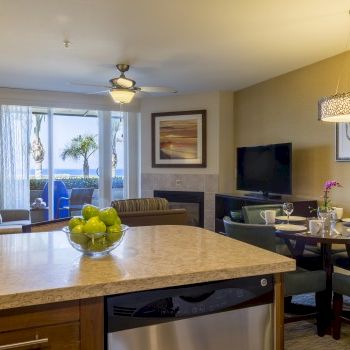 A bright living area with a kitchen island, couch, dining table, wall art, TV, ceiling light, and a view of a balcony through glass doors.