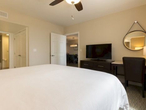 A hotel-style bedroom with a large white bed, flat-screen TV on a dark dresser, round mirror, desk and chair, ceiling fan, and light beige walls. Ends with a period.