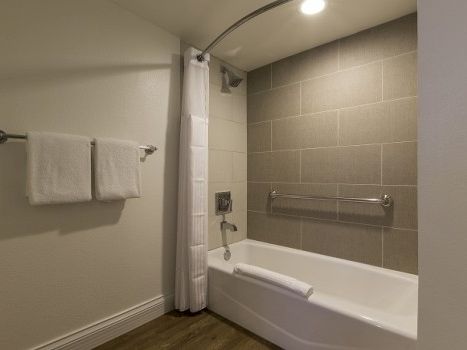 A modern bathroom with a white bathtub, gray tiled wall, shower curtain, stainless grab bar, and two white towels on a rod.
