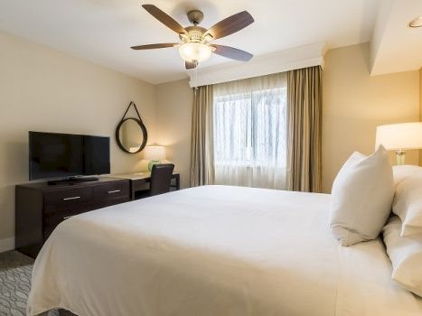 A comfy hotel room with a king bed, bedside lamps, a TV on a dresser, a ceiling fan, a window with drapes, and a circular wall mirror.