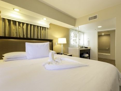 A bright hotel room with a big bed, white linens and folded towel decor, a nightstand lamp, and a small vanity area nearby.