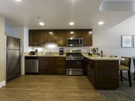 A modern kitchen with dark wood cabinets, stainless steel appliances (refrigerator, stove, microwave), a small island, and a dining area to the right, with wooden flooring and recessed lighting.
