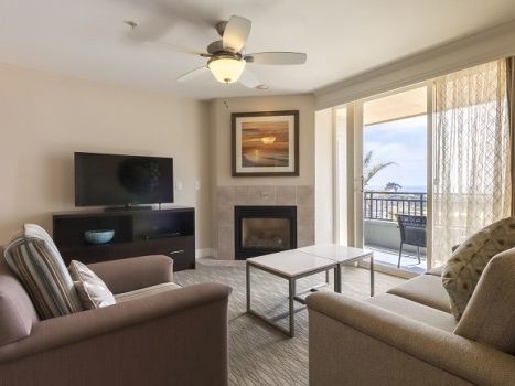 Cozy living room with beige sofas, a wall-mounted TV, a fireplace, a framed art piece, and a glass door opening to a balcony with a sea view, under a ceiling fan.