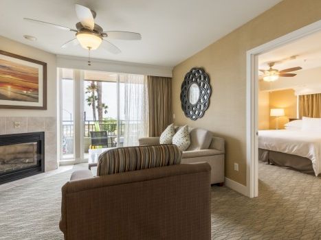 A hotel suite living area with a fireplace, beige walls, a striped sofa, large glass doors to a balcony, a round wall mirror, and a doorway to a bright bedroom.
