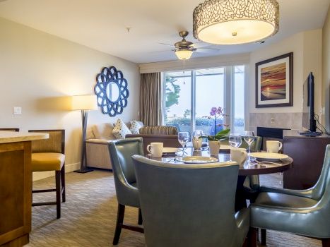 A bright hotel living room with a round dining table, teal chairs, sofa, a TV, large window, decor, and soft lighting.