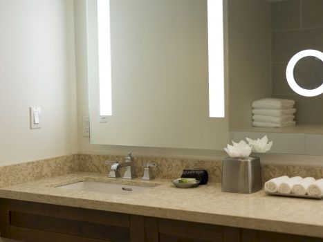 A modern bathroom vanity with a marble countertop, double sink, lit mirrors, stacked towels, and a small ring light in the cabinet.