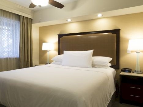 A tidy hotel bedroom with a large bed, white linens, two bedside tables with lamps, a cushioned headboard, and a ceiling fan in warm lighting.