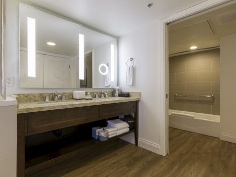 A modern bathroom with a double sink vanity, large mirror with vertical lights, wood-tone cabinet, open shelf, and a separate tiled shower/bathtub area.