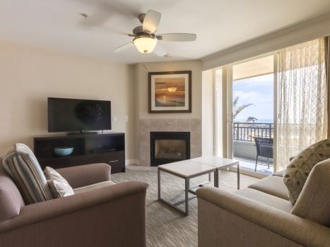 Cozy living room with a sofa and armchairs, fireplace, wall TV, coffee table, ceiling fan, and a balcony with a view, bright and inviting.