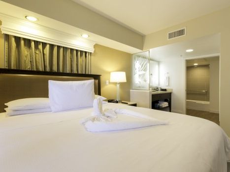 A clean hotel room with a large bed, white linens, a headboard, bedside lamp, and a seating/desk area near a doorway, all in soft lighting.