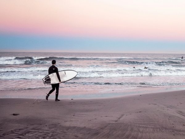 A lone surfer walks along a calm beach at dusk, carrying a surfboard toward the gentle waves, with a moon rising over distant cliffs in the background.