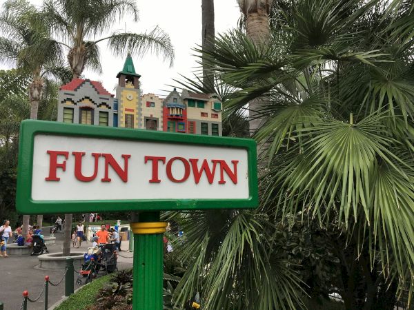 A sign reading &ldquo;FUN TOWN&rdquo; with colorful buildings atop, set among palm trees and people in a themed amusement area.
