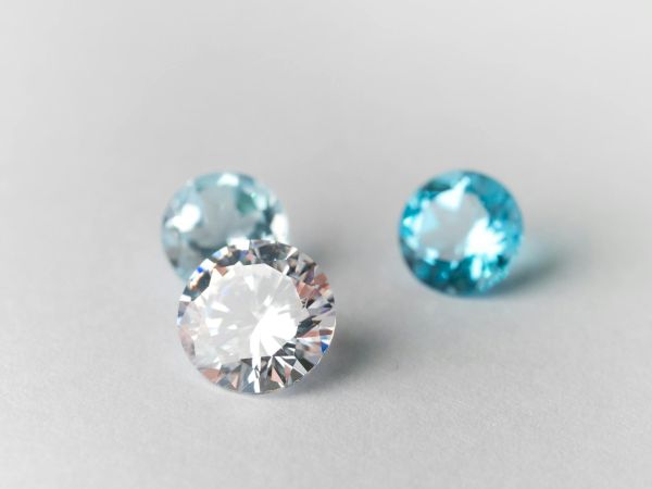 Three sparkling round gemstones on a pale surface: a clear diamond in front, with a light blue stone behind and another teal-blue gem to the right.