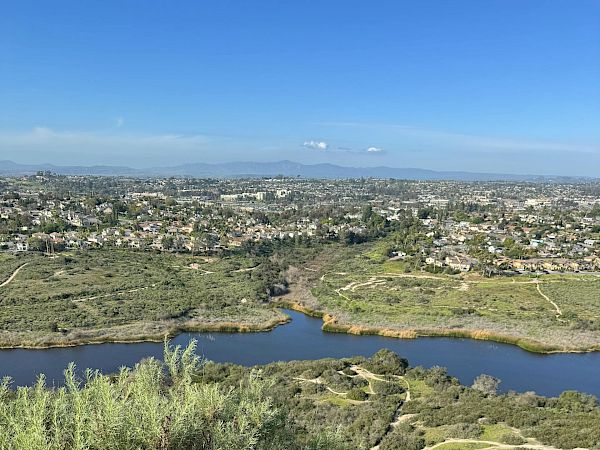 A scenic overlook shows a winding river through scrubland with a distant town and clear blue sky, foreground bushes and a dirt path.