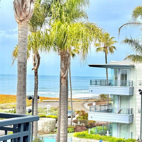 Sunlit beach scene with palm trees, ocean view, and modern balconies from a sunny balcony.