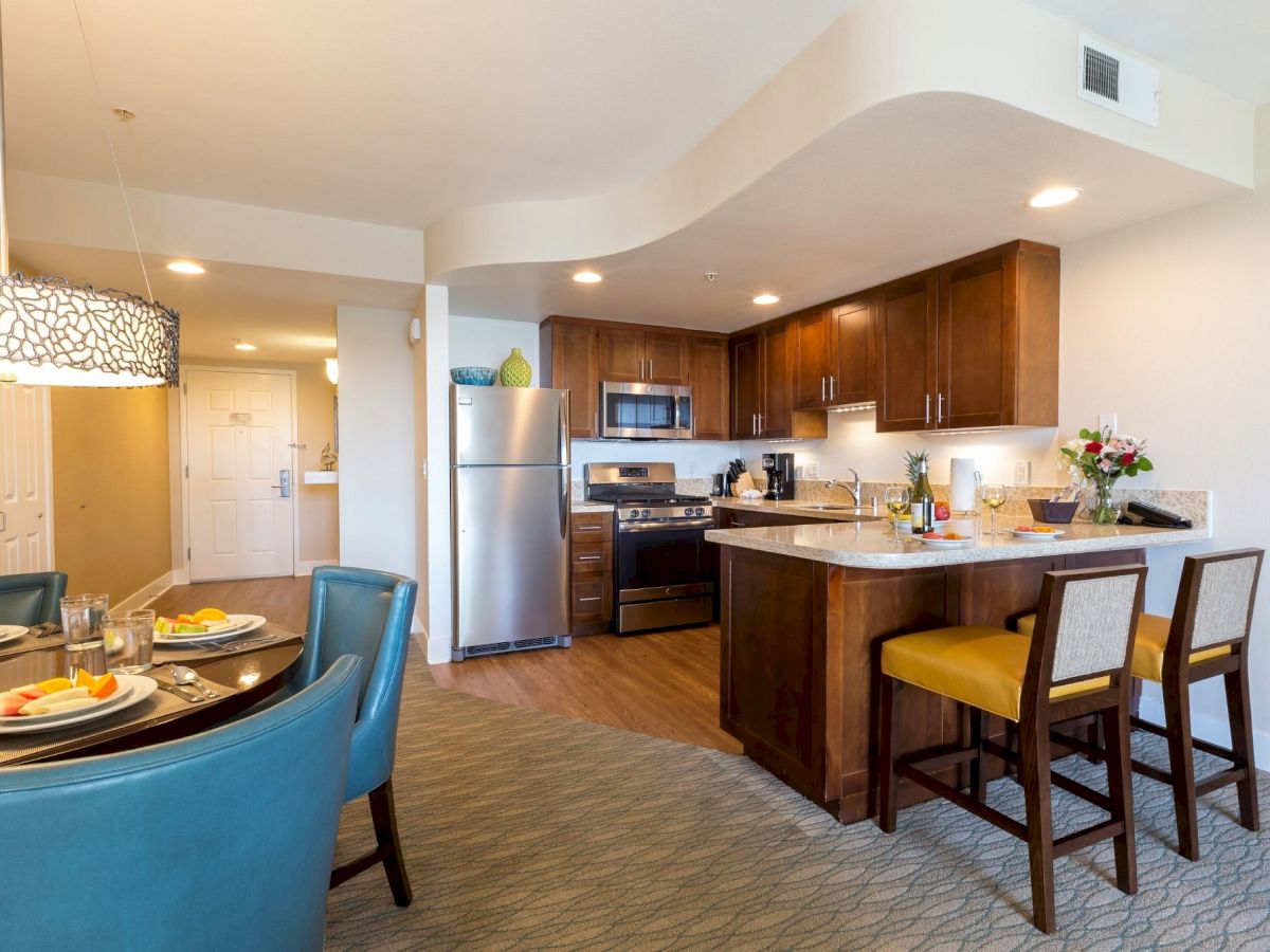 A modern open-concept kitchen and dining area with a dark wood island, stainless appliances, blue chairs at a round dining table, and warm lighting.