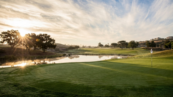 A serene golf course at sunset with a calm pond, green fairways, and a hole flag, bathed in warm light and long shadows.