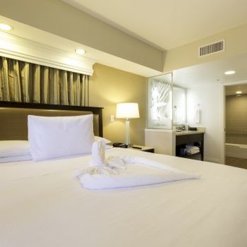 A hotel room with a large bed, white linens, a headboard, bedside lamp, and a mini-sitting area in the back, calm and modern. Ending with a period.