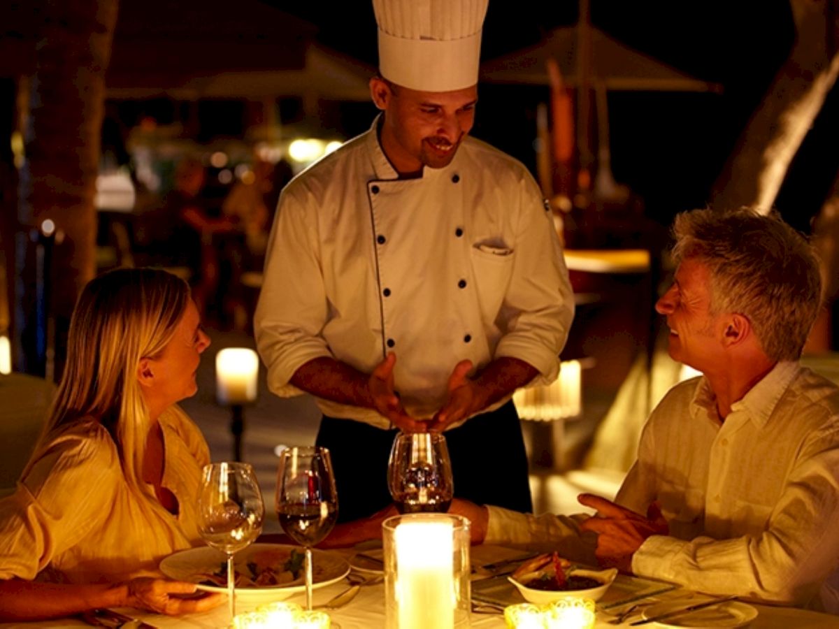 A chef in a white uniform is speaking with a couple at a candlelit table, as a waiter stands between them in a warm outdoor restaurant.